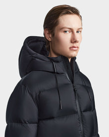 A person with light skin and straight brown hair models the Rudsak DANIEL M - M123510 BLACK weather-resistant winter jacket with a hood, looking slightly to the side against a plain, light background.
