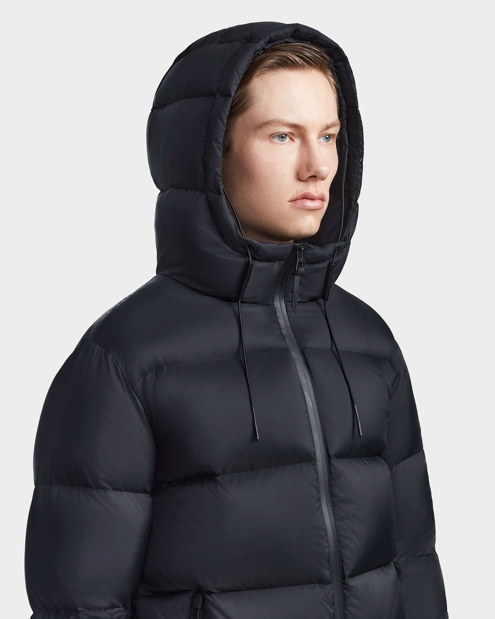 A man wearing the Rudsak DANIEL M - M123510 BLACK hooded down puffer faces forward. The weather-resistant jacket has a zipper and drawstring hood. The background is plain and light-colored.