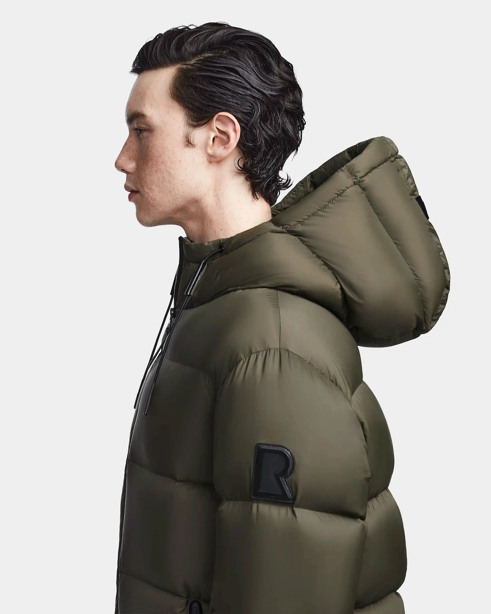 A person with short, dark hair is shown in profile wearing the Rudsak DANIEL M - M123510 Light Olive mens hooded down puffer with a large R patch, designed as a weather-resistant winter jacket, against a plain white background.