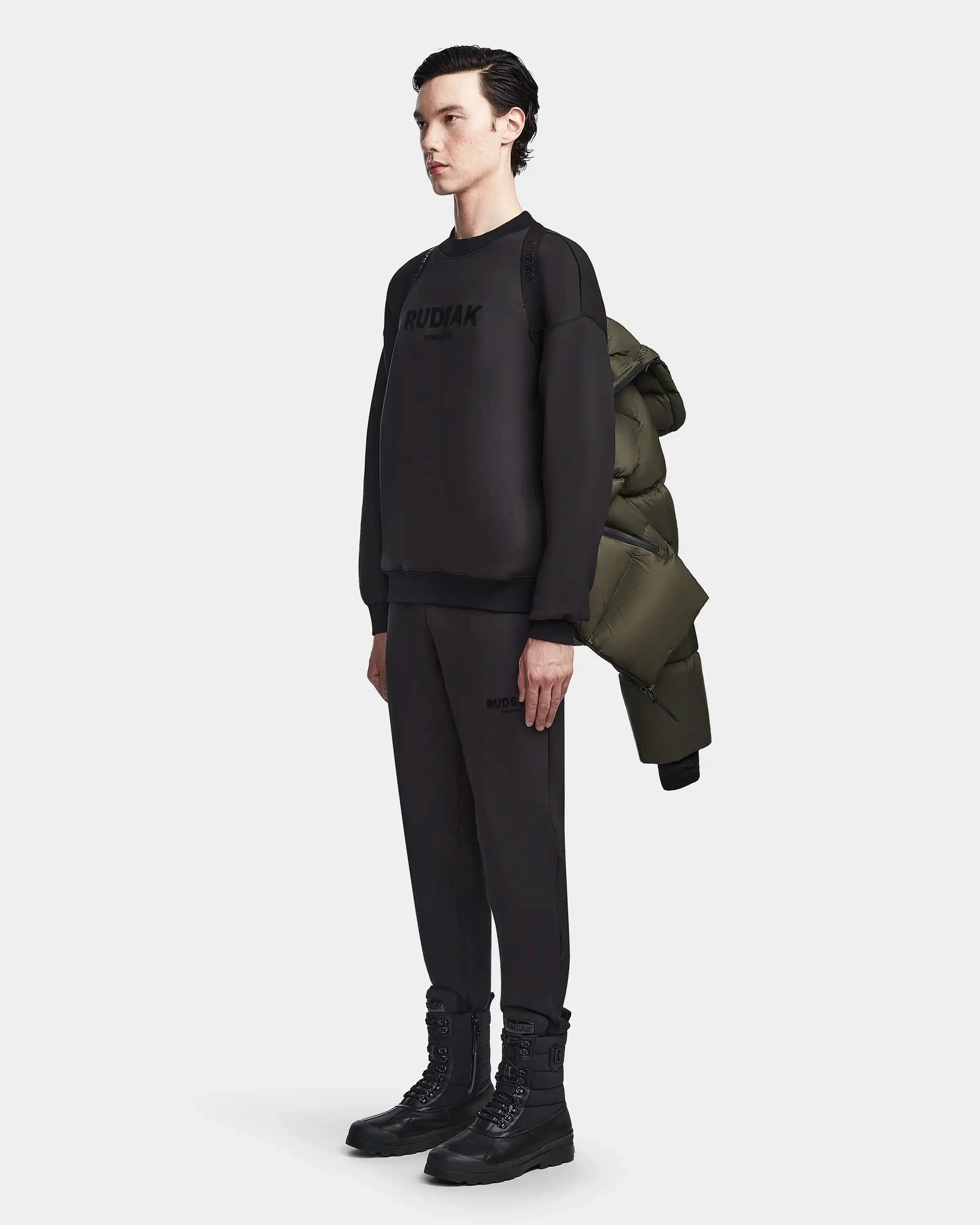 A person stands against a plain background wearing a black outfit and boots, carrying the Rudsak DANIEL M - M123510 Light Olive weather-resistant winter jacket over one shoulder.