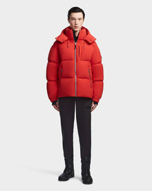 A person faces forward wearing the Rudsak VINCENT M - 6123510 Orange men’s down puffer, paired with black pants and black lace-up boots, set against a plain light background.