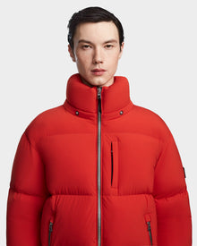 A person with short dark hair wears the Rudsak VINCENT M - 6123510 Orange men's down puffer jacket with a high collar, standing against a plain light gray background.