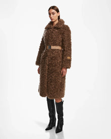 A woman wears the Rudsak KAMILAH - 8124827 Camel faux fur trench coat with a vegan leather belt and tall black boots, posing against a plain white background.