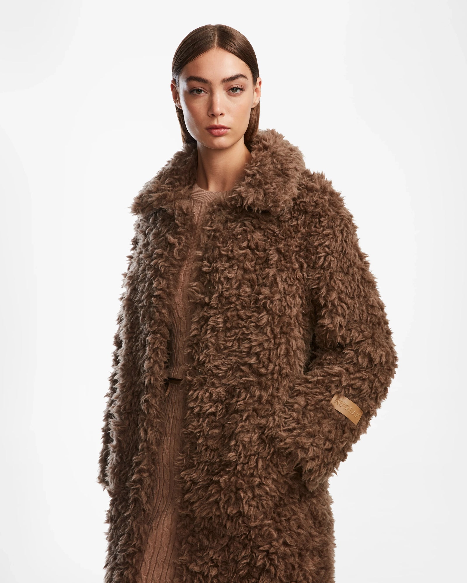 A woman with straight brown hair models the Rudsak KAMILAH - 8124827 camel faux fur trench coat over a matching outfit, standing against a white background and looking at the camera with a neutral expression.