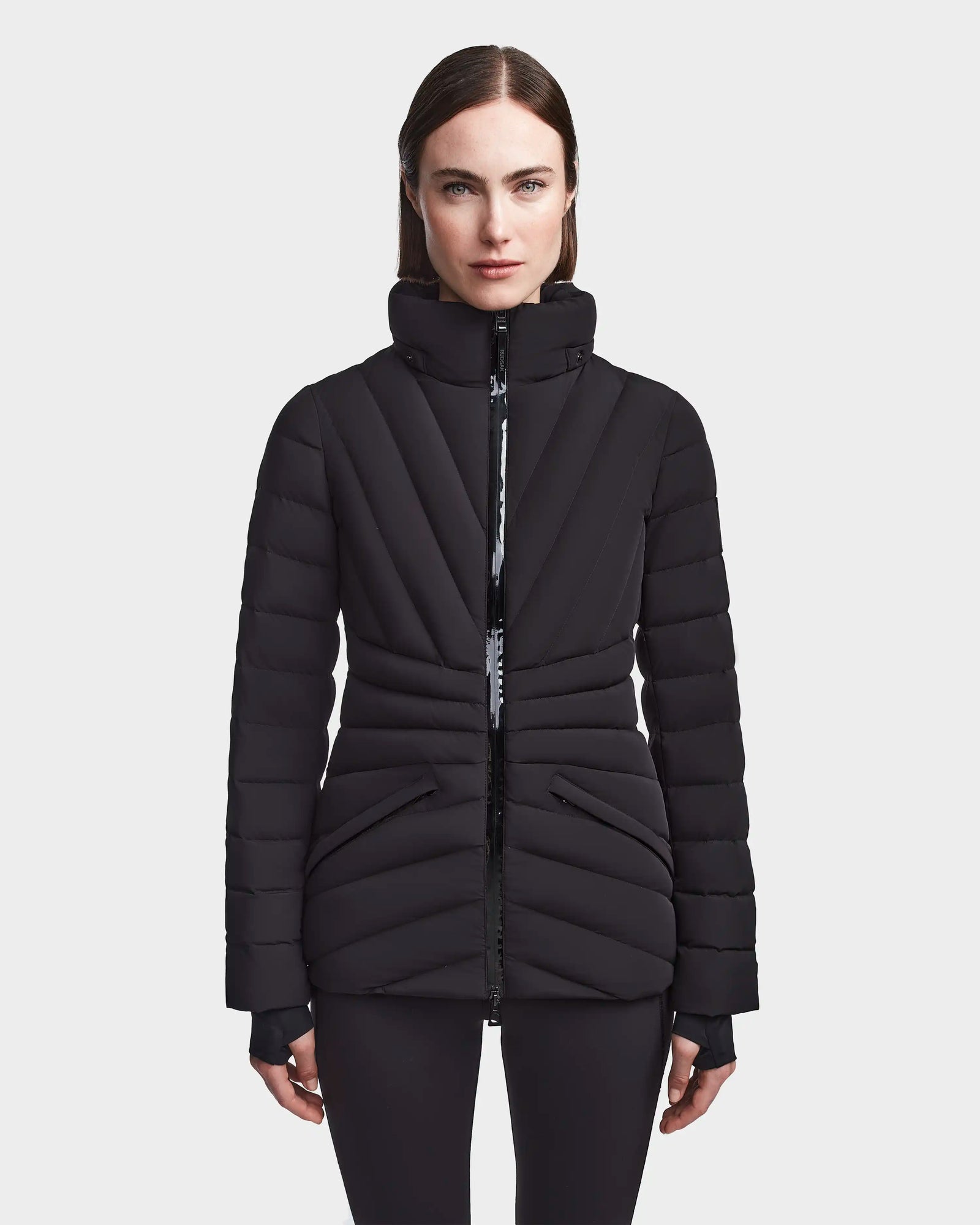 A woman wearing the Rudsak HANNA - 8123562 black winter down jacket with a high collar and horizontal quilting stands facing forward against a plain white background.