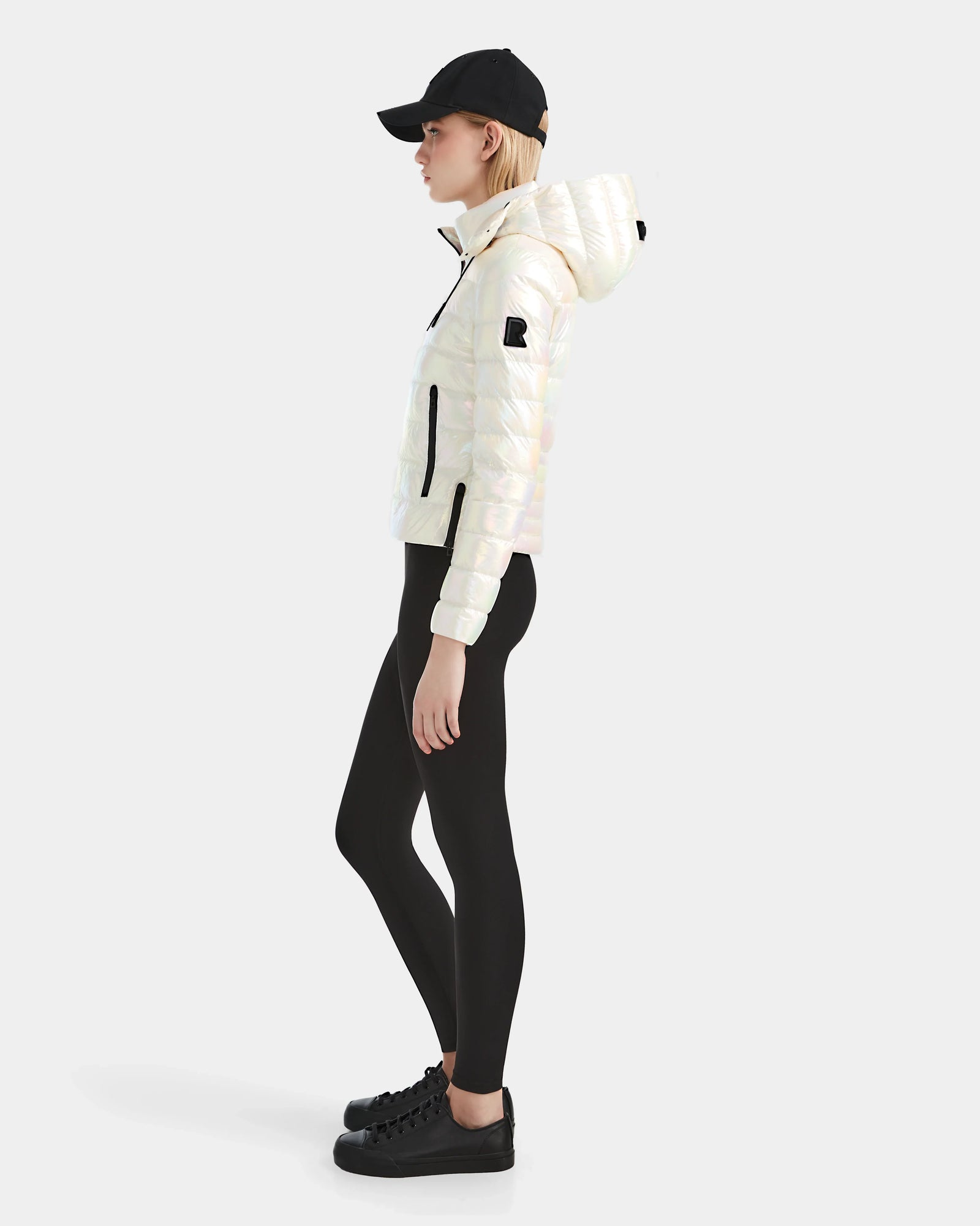 A person in profile wears the Rudsak MELISSA - 8124031 Iridescent Eggshell lightweight down puffer, a black cap, black leggings, and black sneakers against a plain light background.