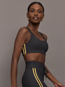 Model is wearing a black scoopneck sports bra with yellow striped sides. 