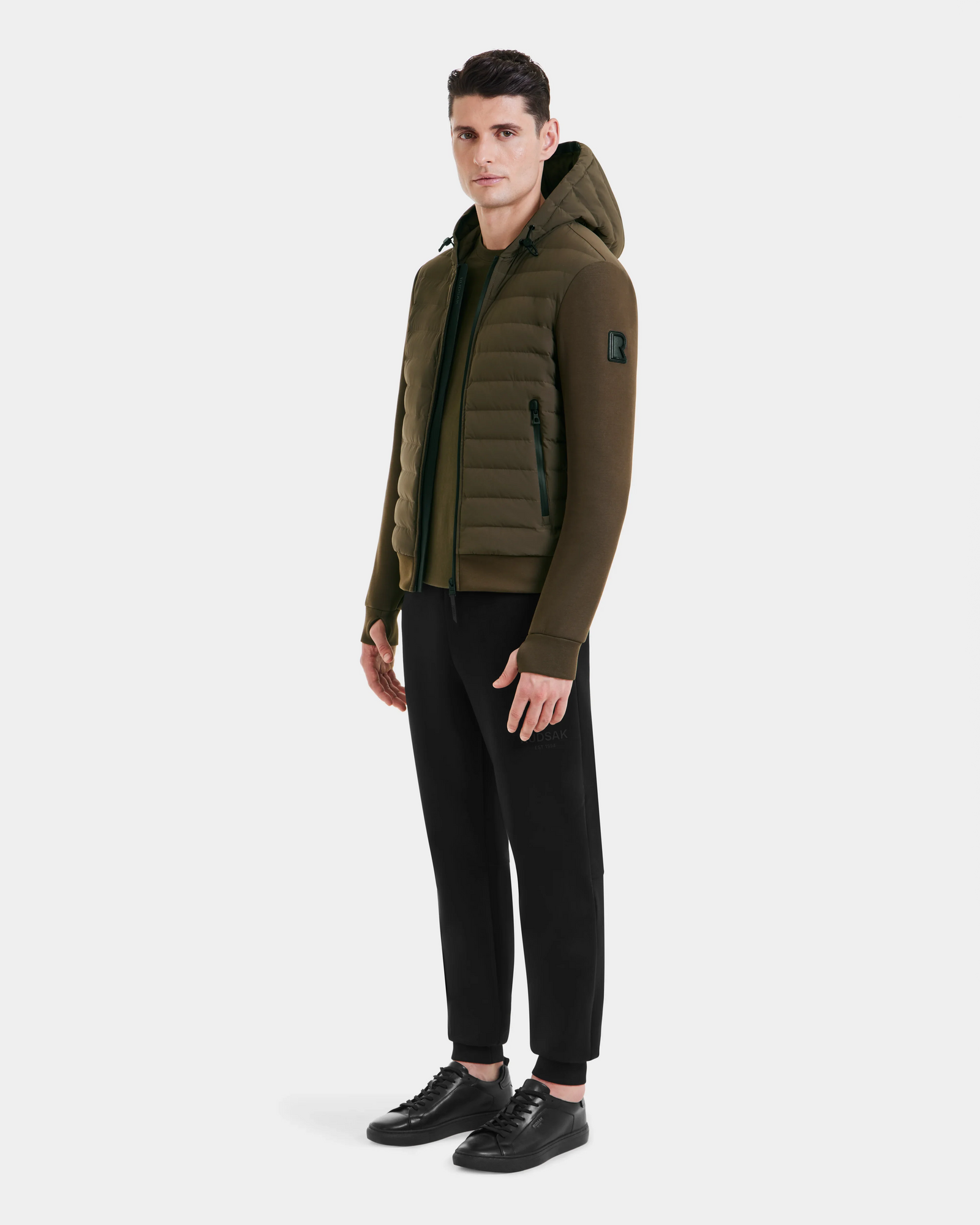 A man stands facing slightly to the side, wearing the Rudsak DEE MAT - 6124028 Army mens lightweight hybrid down jacket, black pants, and black sneakers against a plain white background.