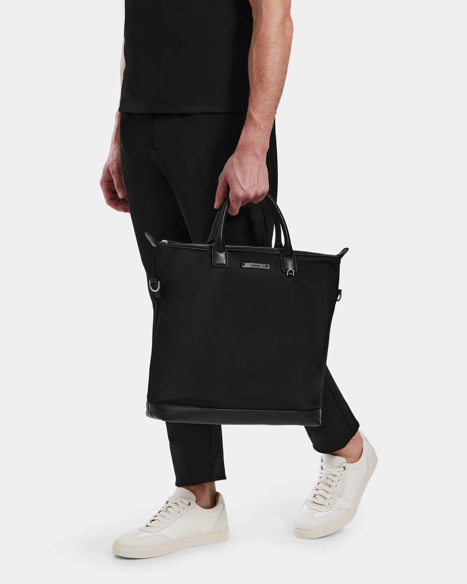 A person in black pants and shirt holds the Rudsak DEXTER - 9324513 Black unisex canvas tote by its handles. Wearing white sneakers, they stand against a plain light background. The person’s face is not visible.