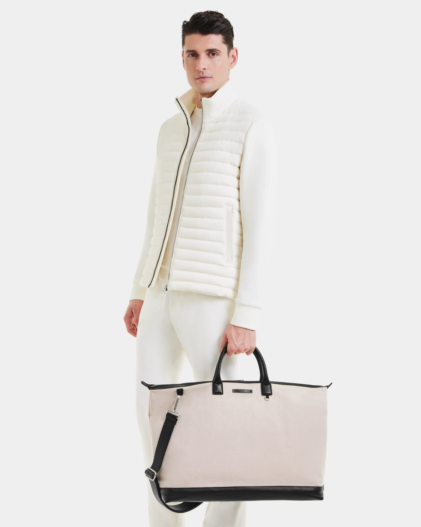 A man in an all-white outfit and quilted vest holds the Rudsak DUBLIN - 9324514 Vanilla unisex weekender tote, featuring handles and a shoulder strap, standing against a plain white background.