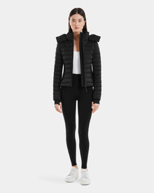 A woman faces forward wearing the Rudsak MARYA M - L123559 Black down puffer jacket, paired with black leggings, a white top, and white sneakers against a plain light background.