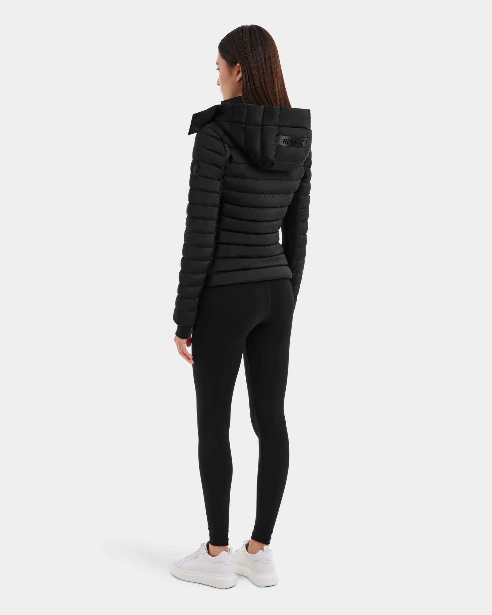 A woman faces away, wearing the Rudsak MARYA M - L123559 Black down puffer with a hood, paired with black leggings and white sneakers, against a plain light background.