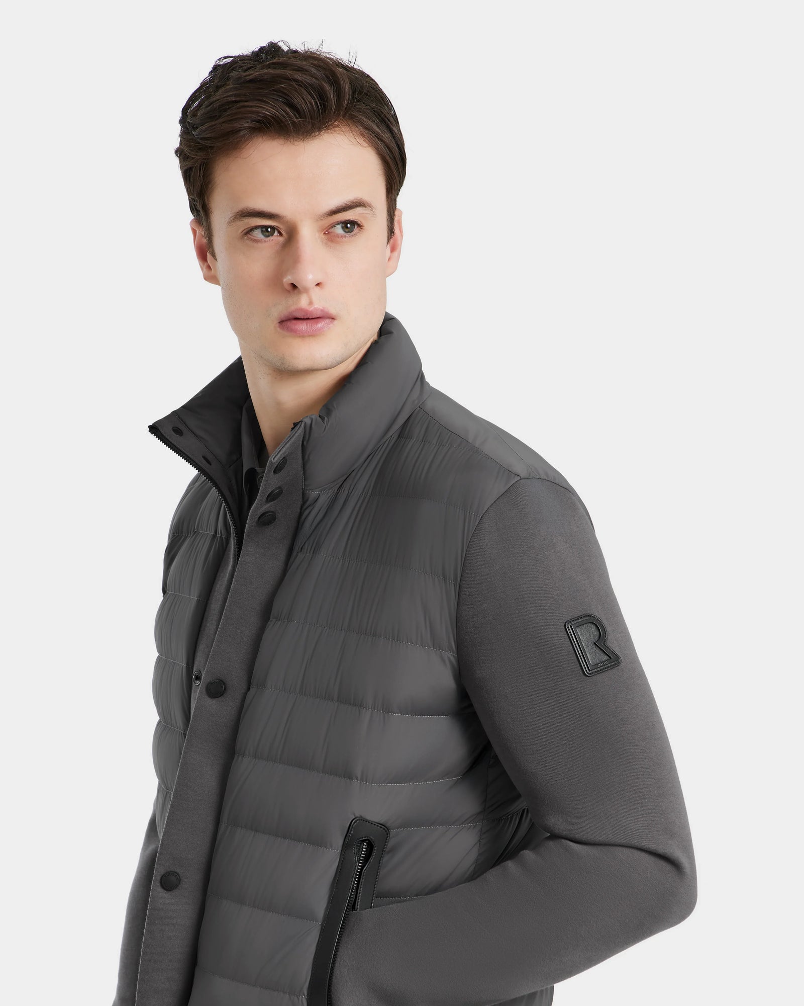 A young man with short brown hair wears the Rudsak RAGAB - 6124577 Grey mens hybrid jacket, featuring lightweight down insulation, a high collar, and black zipper details. He looks to his left against a plain light background.