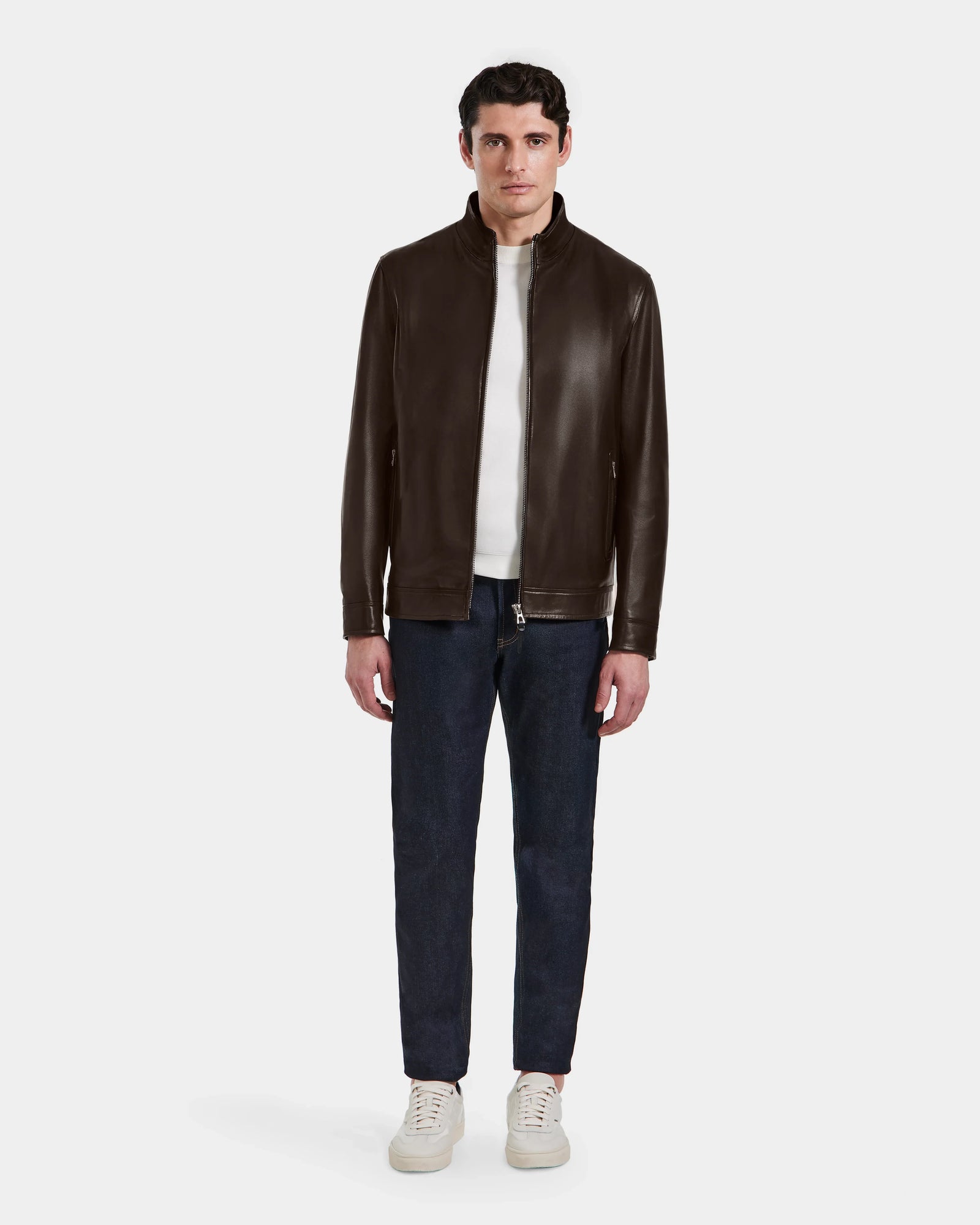 A man faces forward wearing the STEVEN - 6124541 Espresso leather jacket by Rudsak over a white shirt, dark blue jeans, and white sneakers, set against a plain white background.