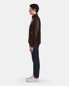 A man stands in profile against a white background, wearing the Rudsak STEVEN - 6124541 Espresso leather jacket, paired with dark blue jeans and white sneakers.