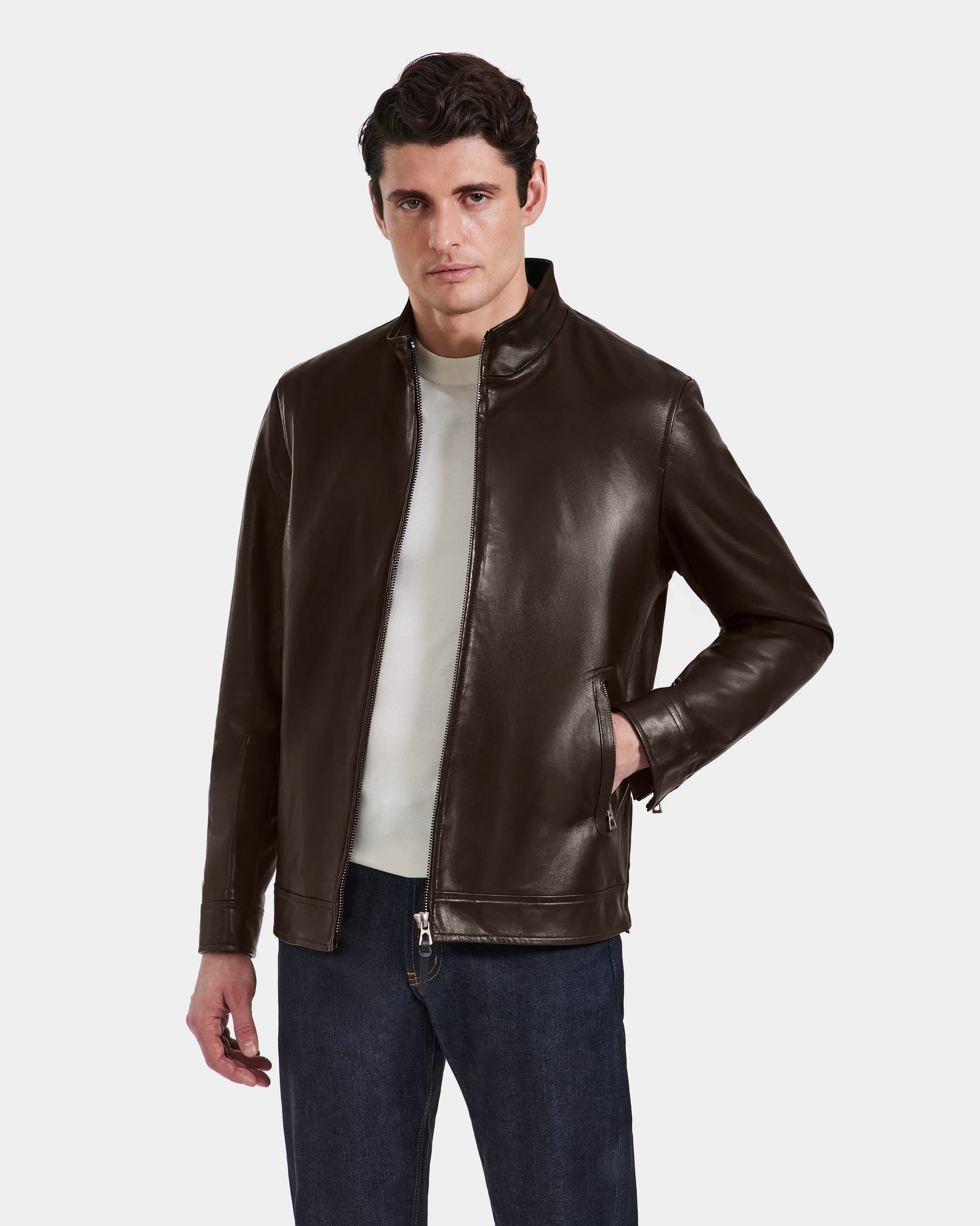 A man wears the Rudsak STEVEN - 6124541 Espresso leather jacket over a white shirt and blue jeans, standing against a plain light gray background with one hand in his pocket and a neutral expression.