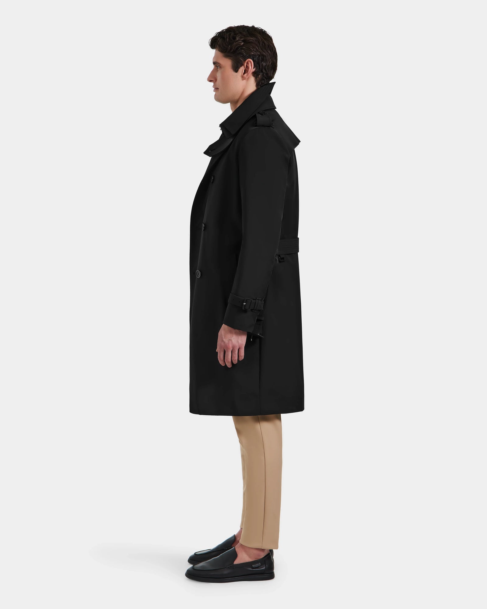 A man stands in profile against a plain background, wearing Rudsaks TIMOTHY - 6124589 Black knee-length trench coat, paired with beige pants and black loafers.