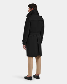 A man stands facing away, wearing the Rudsak TIMOTHY - 6124589 Black knee-length trench coat, beige pants, and black shoes against a plain light background.