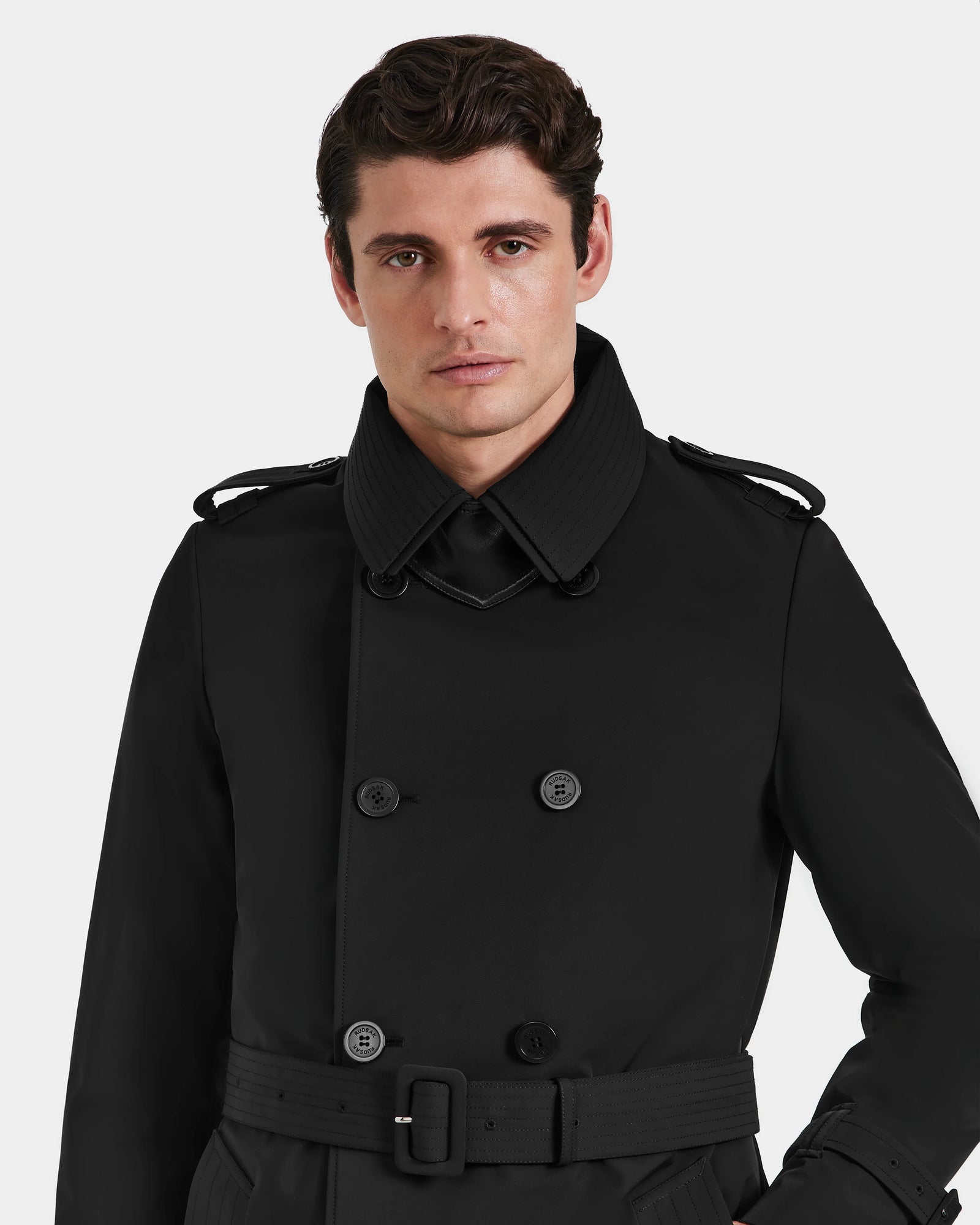 A man with short brown hair wears the Rudsak TIMOTHY - 6124589 black mens trench coat, featuring a belt and shoulder epaulets, standing against a plain white background.