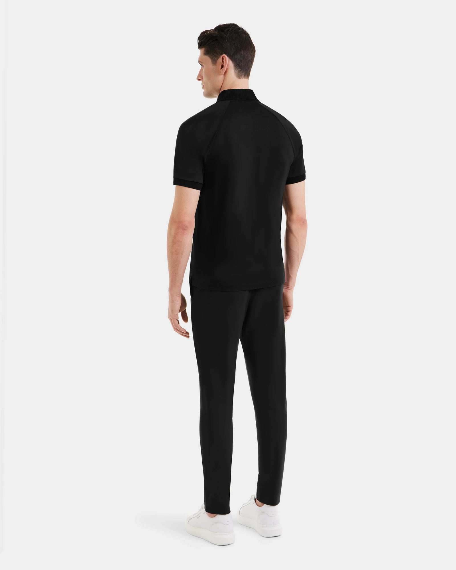 A man faces away, wearing the Rudsak WALTER - 6524531 Black/Black men’s short sleeve polo with matching black pants and white sneakers against a plain light background.