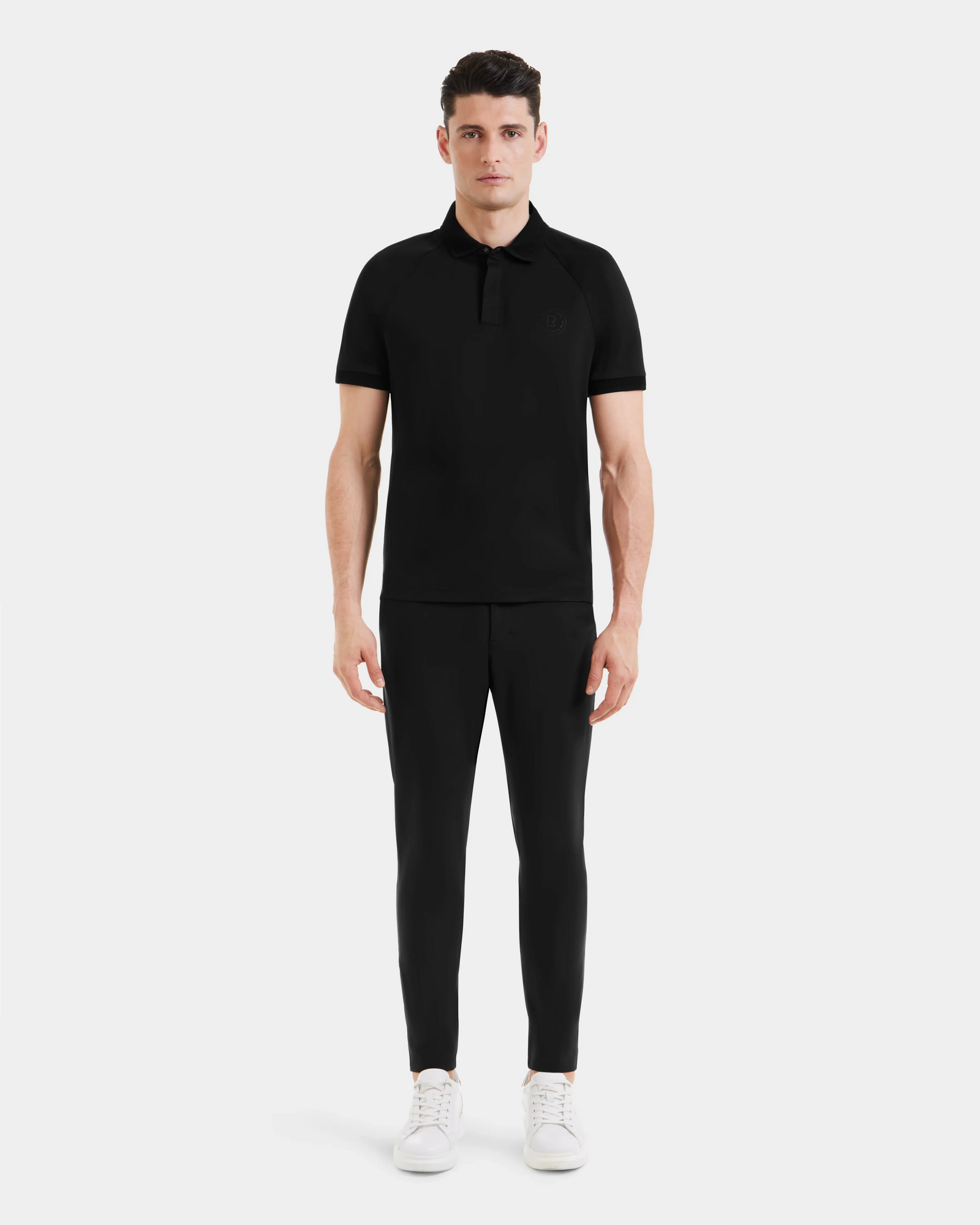 A man stands facing forward wearing the WALTER - 6524531 Black/Black polo by Rudsak, paired with black pants and white sneakers against a plain light background.