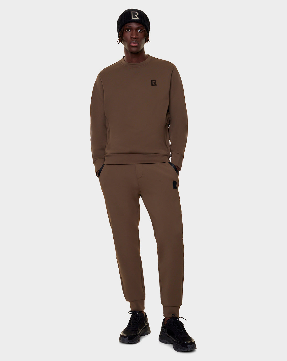A person stands facing forward in Rudsak’s DEVAN - 9522510 ARMY MEN brown relaxed-fit sweater and joggers lounge set with black sneakers and a black beanie, each featuring a small R logo, against a plain light background.