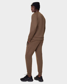 Wearing the Rudsak DEVAN - 9522510 ARMY MEN relaxed fit unisex crewneck sweater and matching jogger pants, paired with black sneakers and a black knit beanie, a person stands facing away, looking over their shoulder against a light gray background.