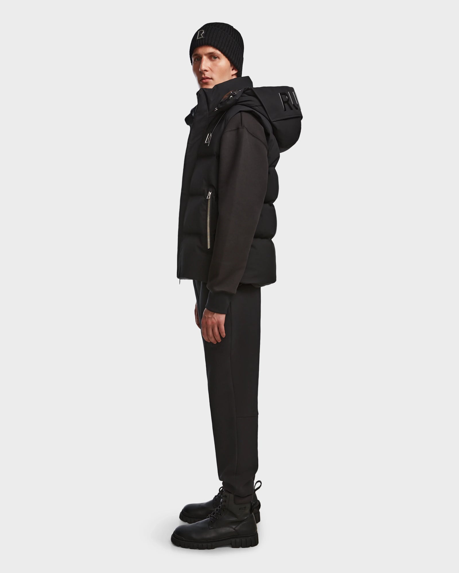 A man stands in profile against a white background wearing the Rudsak REN M - 9123512 Black M weather-resistant beanie, RDS-certified down puffer vest with hood, jacket, fitted pants, and lace-up boots.