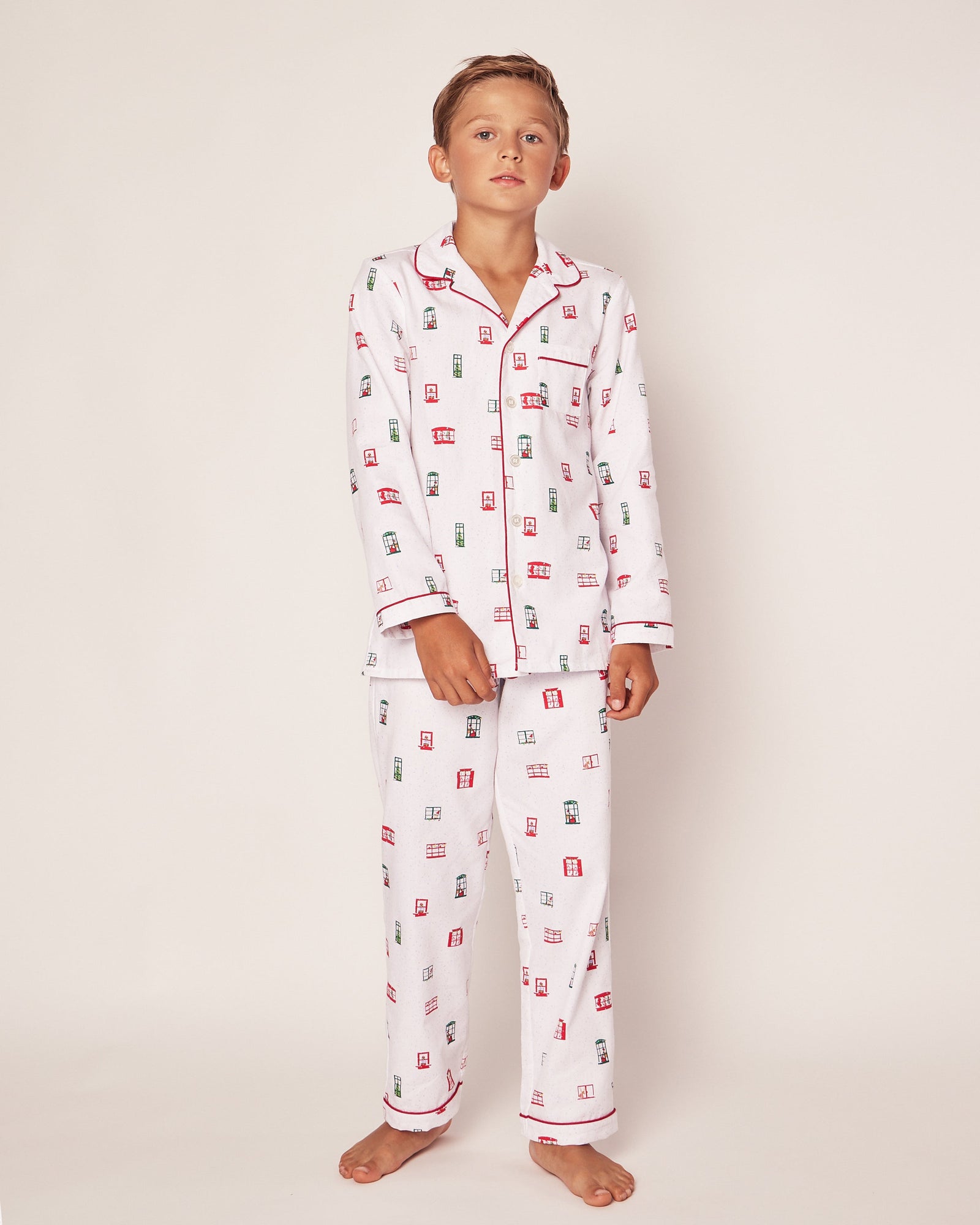 A young boy stands against a plain background in the luxurious Petite Plume Sleepwear Set, featuring a red and green Night Before Christmas print. The pajamas are part of the Maisonette collaboration and include red trim along the collar and sleeves. Hes barefoot, looking relaxed at the camera.
