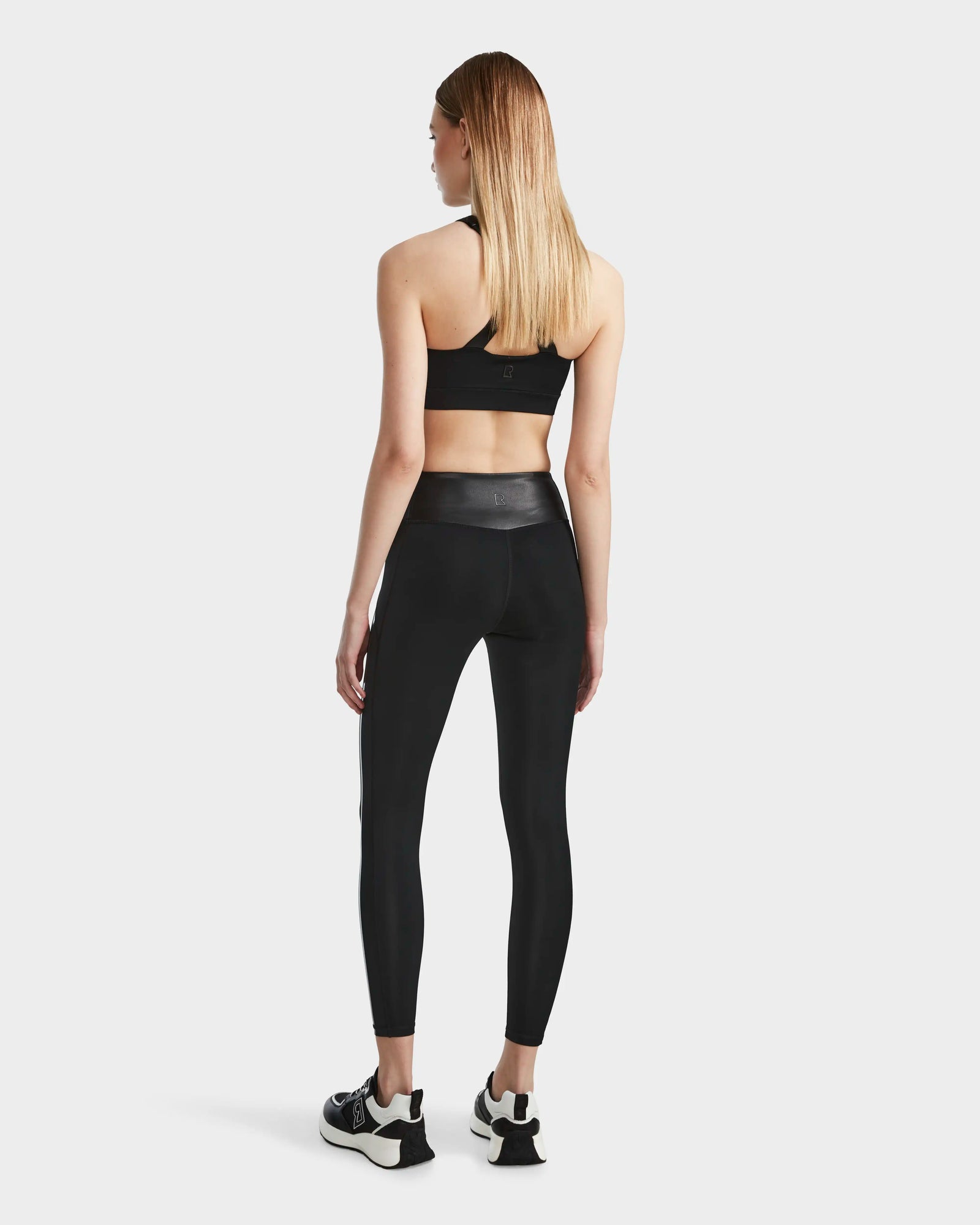 A woman with straight, light brown hair wears Rudsaks THEA - 8523003 BLACK high-rise leggings with a vegan leather waistband and a black sports bra, facing away from the camera against a plain white background.