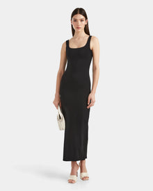 A woman wears the Rudsak WANDA - 8524035 Black fitted sleeveless maxi dress with stretch performance fabric, paired with white high-heeled sandals and a small white woven handbag, against a plain white backdrop.