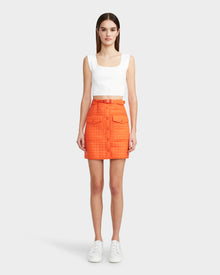 A woman faces forward in a Rudsak SABINE - 8524063 Orange Sorbet quilted mini skirt with buttons, pockets, and a matching belt, paired with a sleeveless white top and white sneakers against a light gray background.