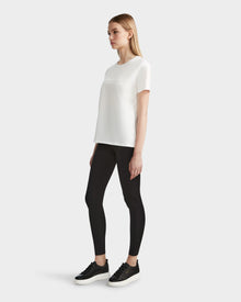 A woman stands against a plain background wearing the Rudsak AMINA - 8524012 Cotton White womens short sleeve t-shirt, black leggings, and black sneakers. She is facing right with her arms at her sides.