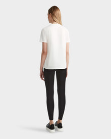 A person with long, straight blonde hair stands facing away, wearing the Rudsak AMINA - 8524012 Cotton White womens short sleeve t-shirt, paired with black leggings and black sneakers with white soles against a plain light background.