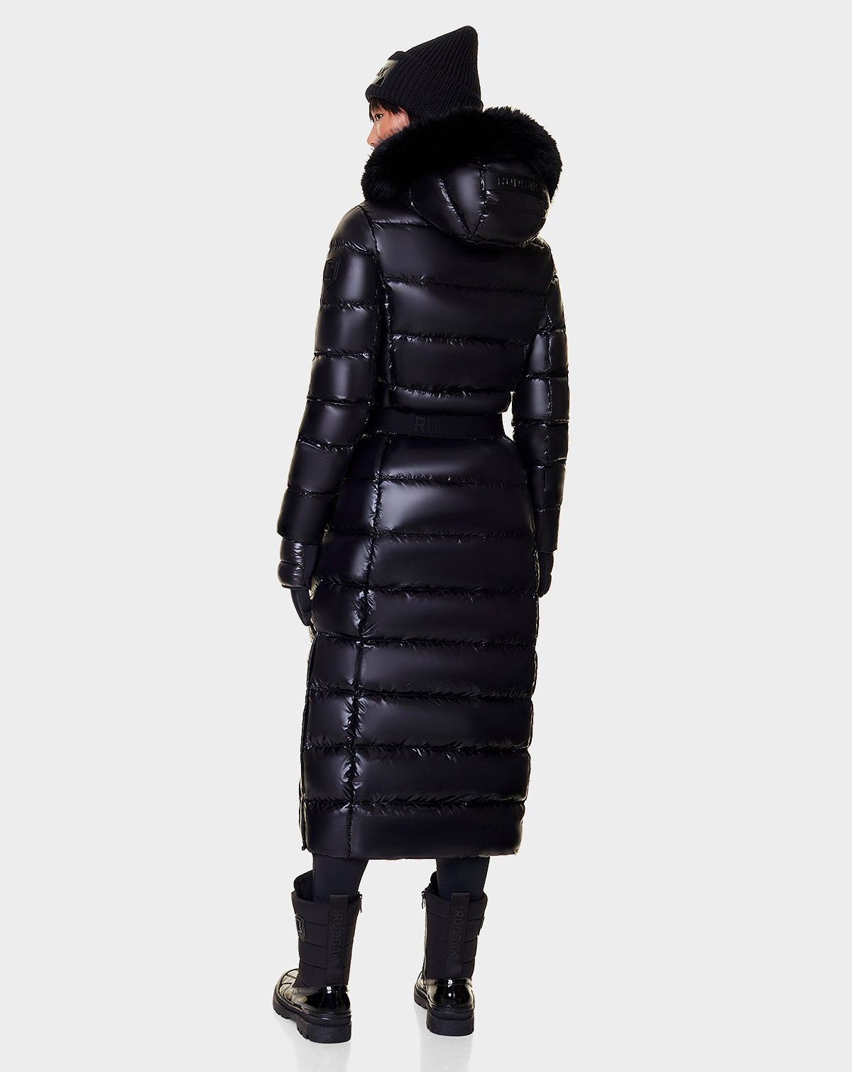 A person is seen from the back wearing the Rudsak ELSIE - L121525 BLACK women’s maxi down puffer with a recycled semi-gloss exterior, fur-trimmed hood, and shiny black finish, paired with black gloves, pants, shoes, and knit hat against a light background.