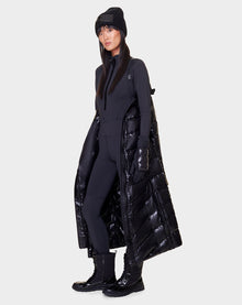 A woman faces forward in a modern, all-black winter look, wearing the Rudsak ELSIE - L121525 BLACK maxi down puffer with a recycled semi-gloss exterior. The stylish coat exudes warmth against a plain background.