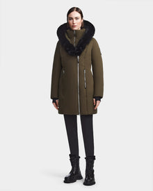 A woman stands facing forward in a Rudsak MAIA M - 8123582 Army womens down parka with a large black fur-trimmed collar, paired with black leggings and lace-up boots against a plain white background.