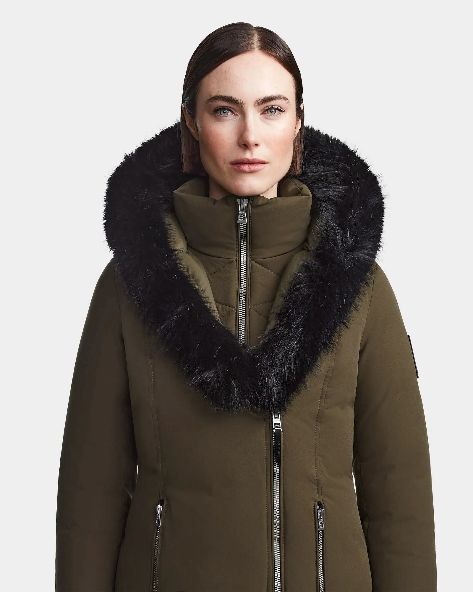 A woman with straight brown hair models the Rudsak MAIA M - 8123582 Army womens down parka in olive green, featuring a large black faux fur-trimmed hood and silver zippers, standing against a plain white background.