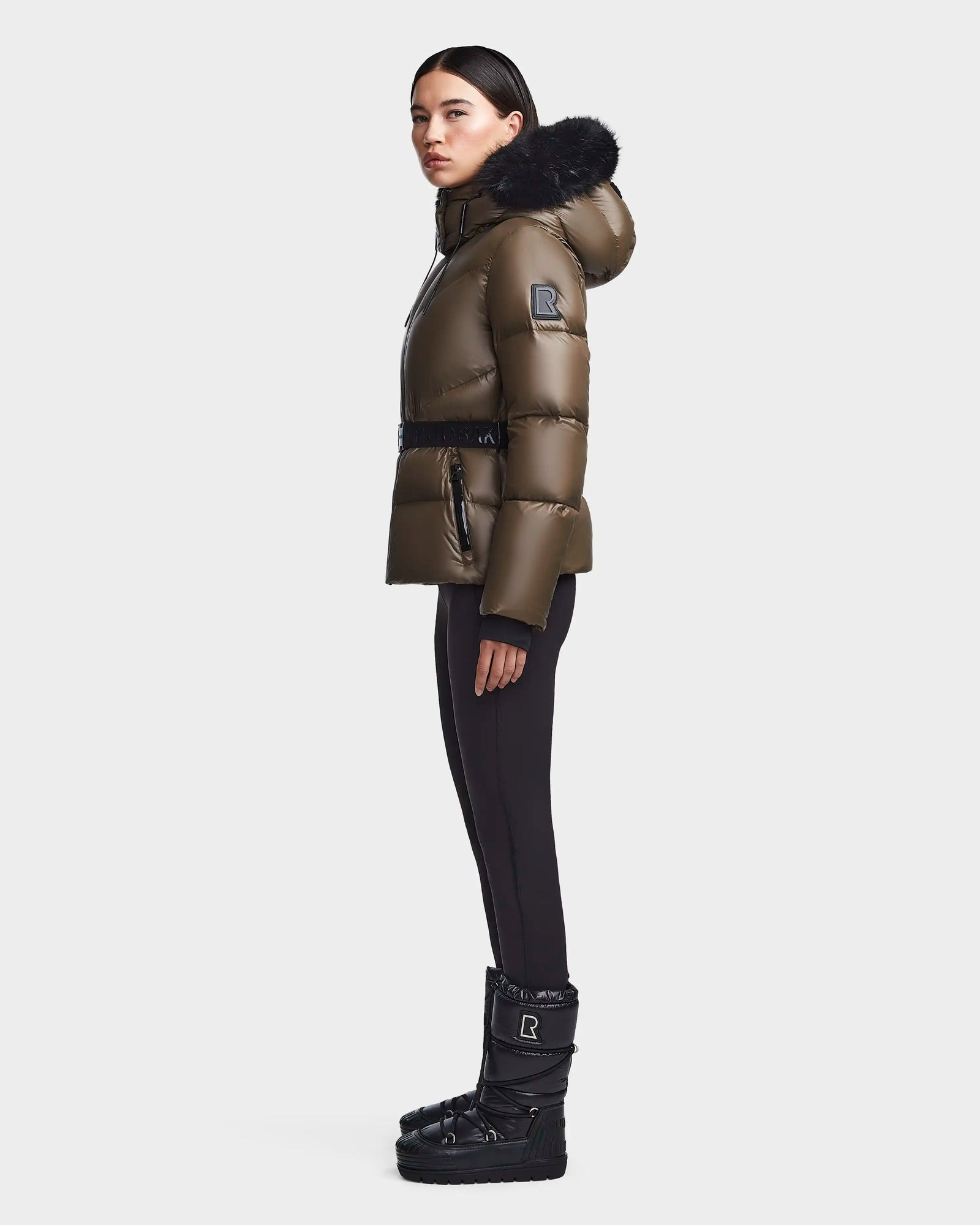 A woman in profile wears the Rudsak CHRISTELLE S - L123524 Light Olive chevron quilted down puffer with black fur hood, black leggings, and winter boots against a plain light gray background.