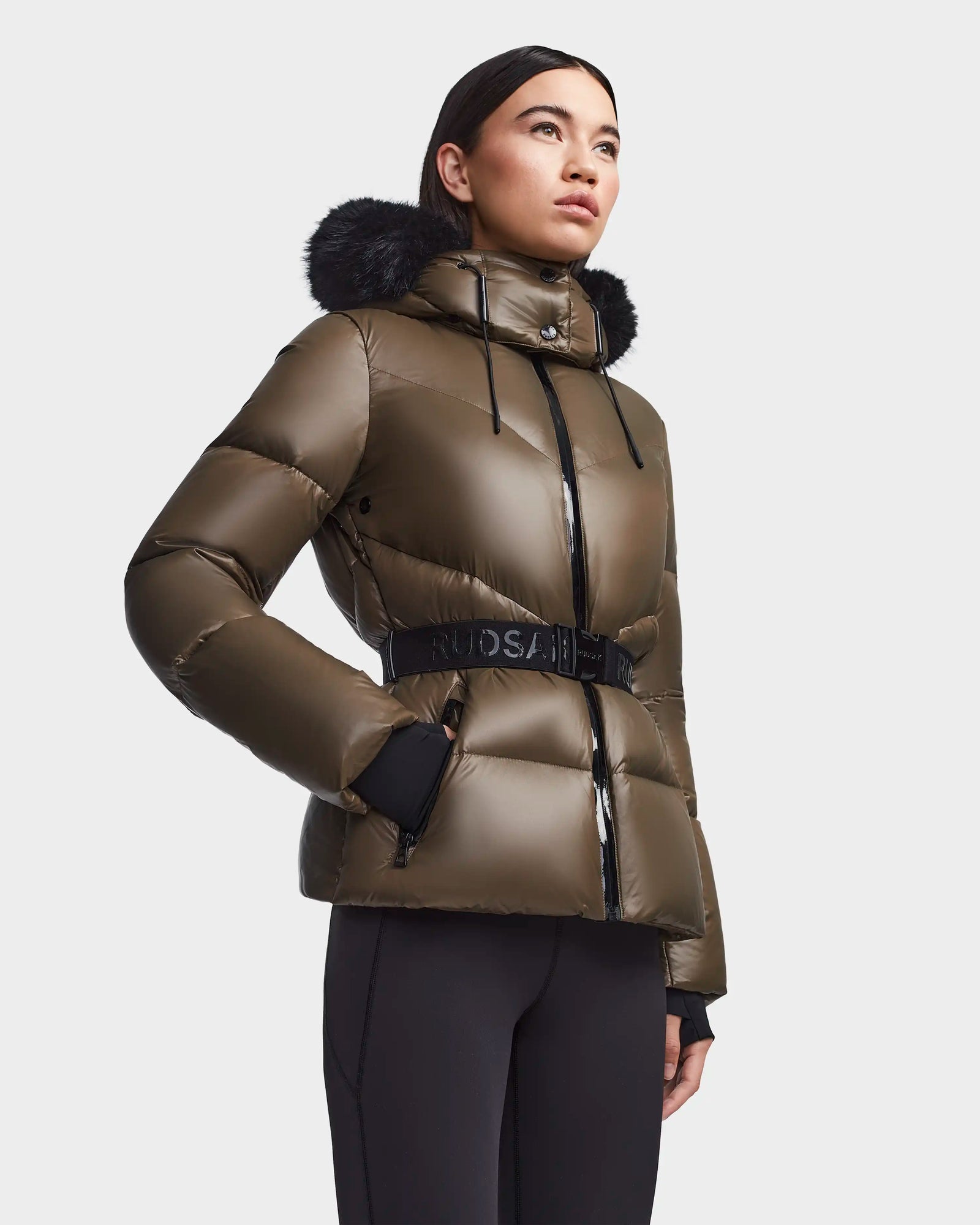 A person wearing the Rudsak CHRISTELLE S - L123524 Light Olive women's quilted down puffer with a black fur-trimmed hood stands against a plain background, looking slightly upwards.