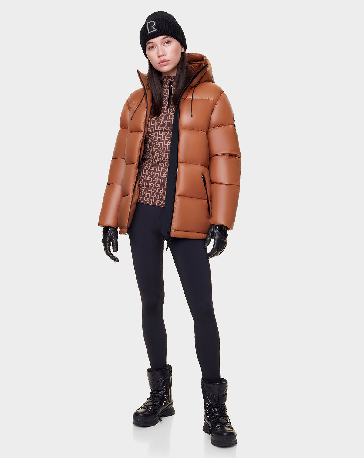 A woman stands against a white background wearing the Rudsak EVIE - L122517 DARK CAMEL down puffer for women with waterproof zippers, black leggings, winter boots, gloves, a patterned top, and a black beanie.