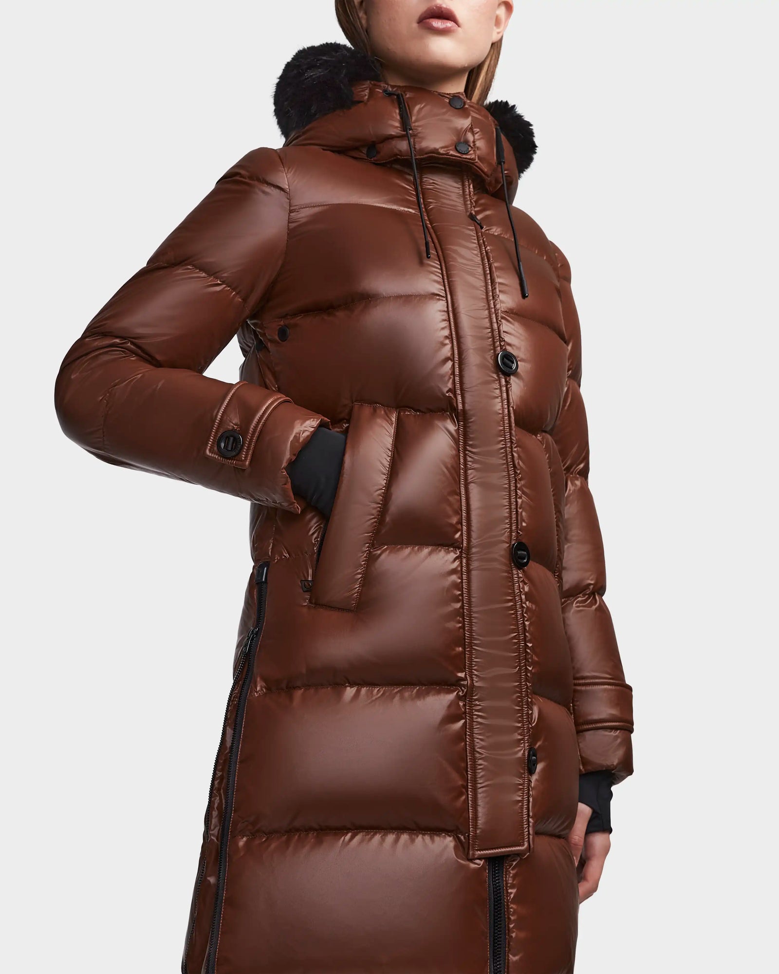A woman wearing the Rudsak MEYA S - L123521 Cohiba shiny brown down puffer coat with removable hood and black faux fur trim, large buttons, and zip details is shown from mid-thigh up against a plain background.