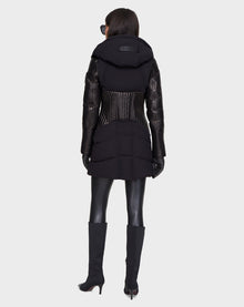 A person wearing the Rudsak SHAUNA - 8121509 BLACK quilted down puffer coat with leather sleeves, black gloves, leggings, and knee-high heeled boots stands facing away against a plain light background.