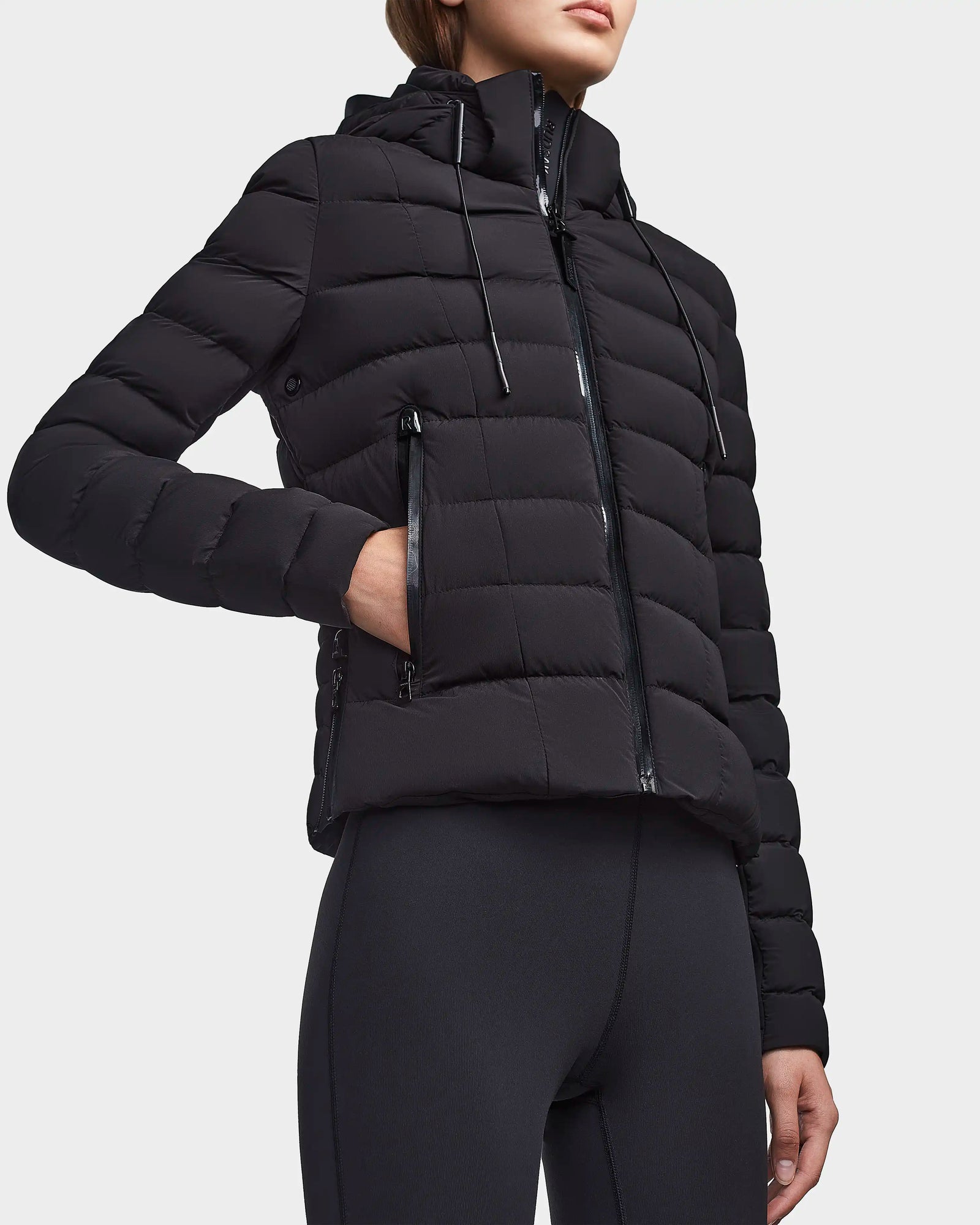 Someone stands against a plain light background wearing the RUDSAK MELISSA - 8122551 BLACK fitted, weatherproof down jacket with a hood and zippered pockets, paired with black leggings.