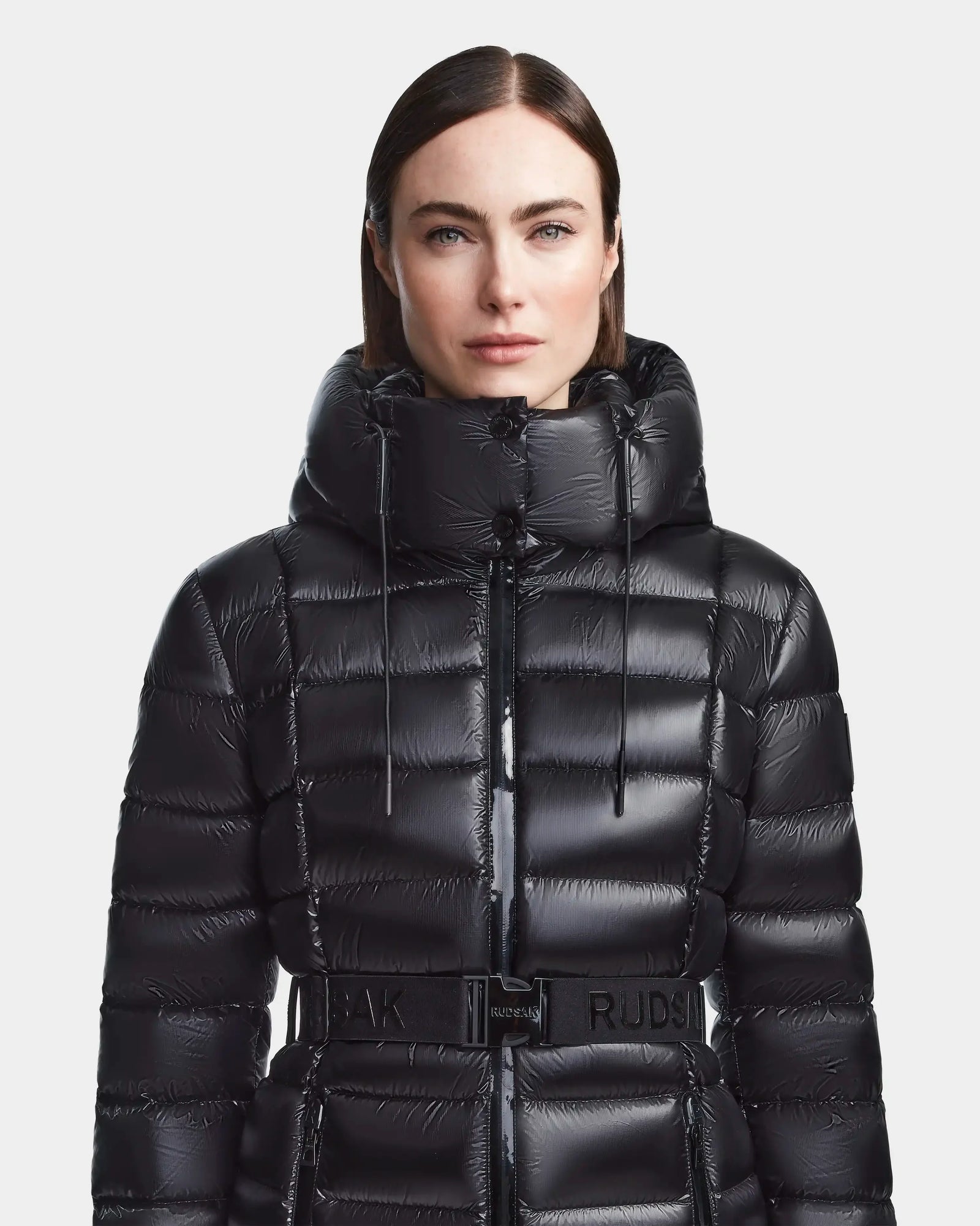 A woman with straight dark hair models the Rudsak FRIDA S - L123513 Black, a shiny black women's mid-length down jacket with a high hood and belted waist, standing against a plain white background.