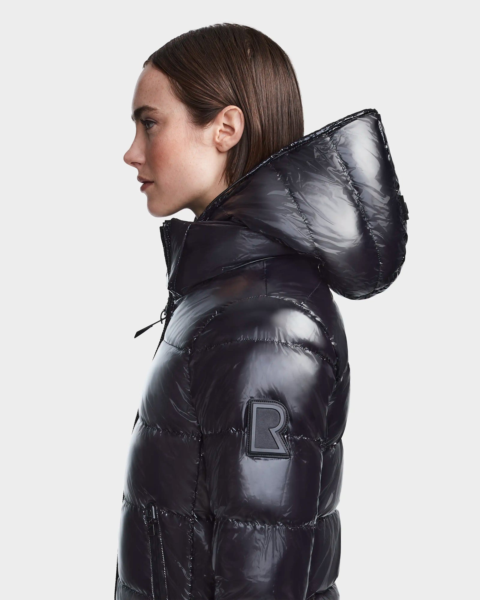 A woman in profile wears the RUDSAK MARNIE LONG - 8122519 black puffer jacket with a hood and an R patch on the sleeve, standing against a plain light background. This jacket blends sleek style and cozy warmth for everyday versatility.