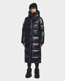 A person stands facing forward against a plain white background, wearing the Rudsak RITA L-S - L123526 Black women's maxi-length down puffer coat, paired with a black knit beanie, black pants, and black shoes.