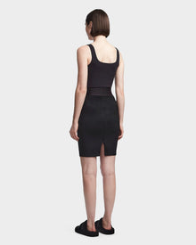 A woman with short brown hair faces away, wearing a sleeveless black top and the CHEYNA - 8523514 Black faux suede pencil skirt by Rudsak, paired with black slip-on sandals against a plain light gray background.