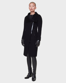 Wearing RUDSAK’s CHAY - 8122506 BLACK long, double-faced wool coat with black gloves, leggings, and ankle boots, a person with a buzz cut poses against a plain white background.