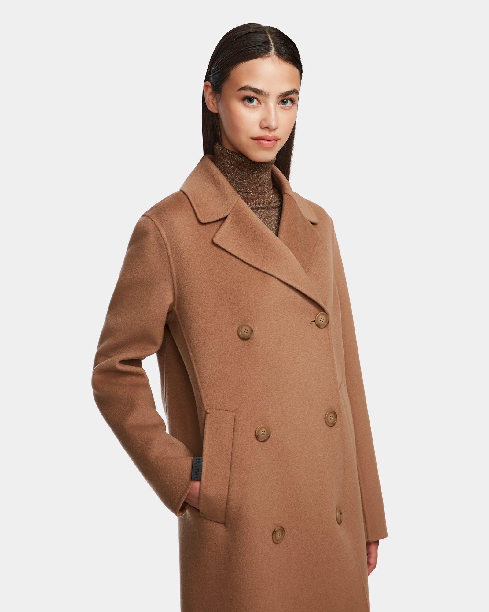 A woman with straight dark hair wears the Rudsak LAYLAH - 8124699 Camel knee-length coat and a brown turtleneck, posing against a plain white background with one hand in her coat pocket.